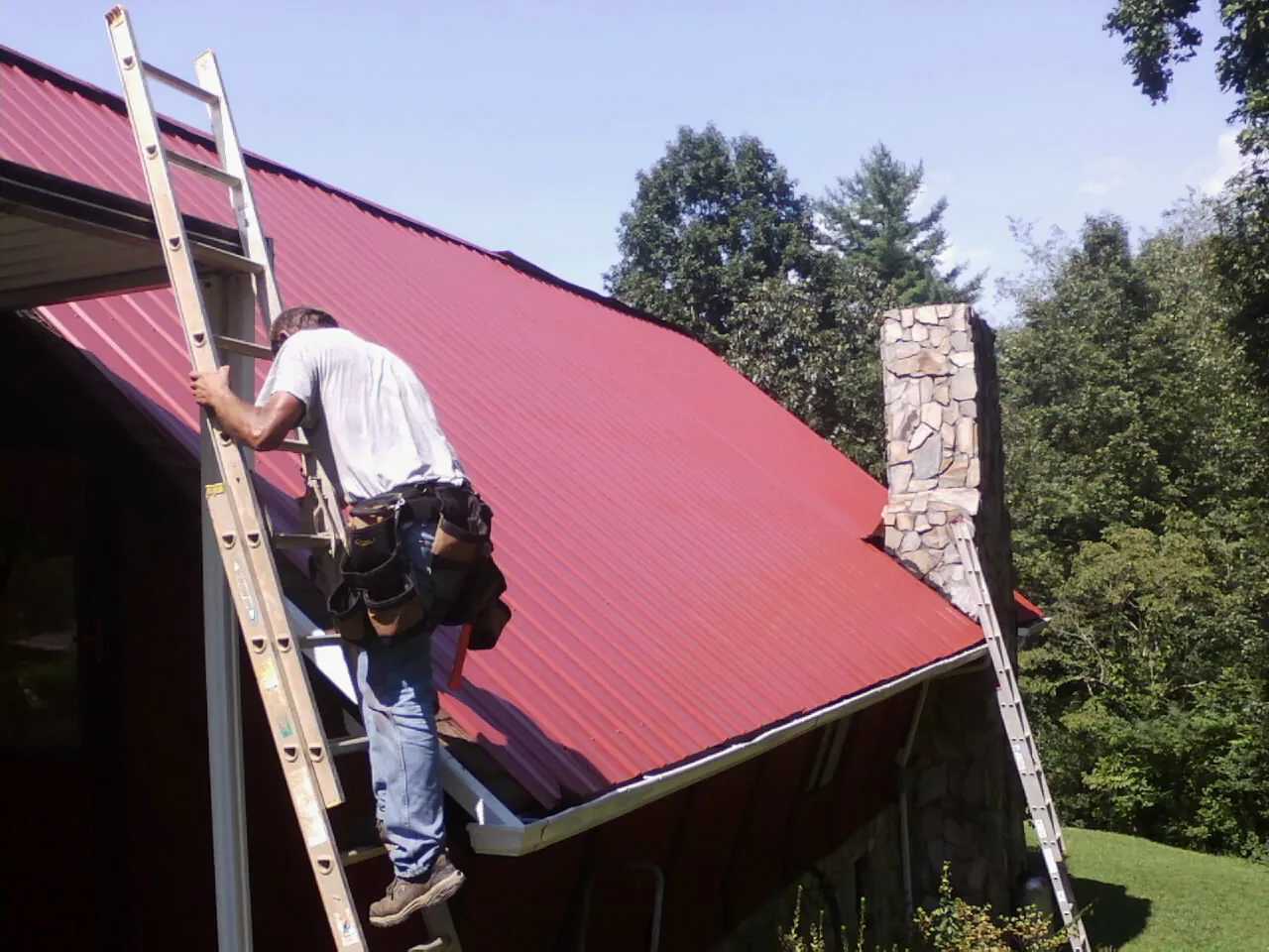 Roofer on a ladder beside a red metal roof and stone chimney — Soffit & Fascia Repair by Roofing Brothers in Nelsonia
